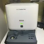 Das Bild zeigt ein Gerät der Marke IDEXX, Modell Catalyst One. Es hat eine weiße Vorderseite und eine graue Ablage. An der Vorderseite befindet sich ein grünes Licht.