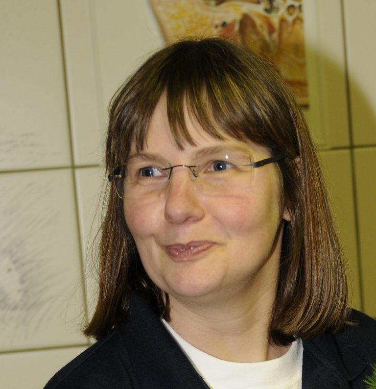 Eine Person mit braunen Haaren und Brille trägt eine dunkle Kleidung. Im Hintergrund sind helle Fliesen und Zeichnungen an der Wand sichtbar.