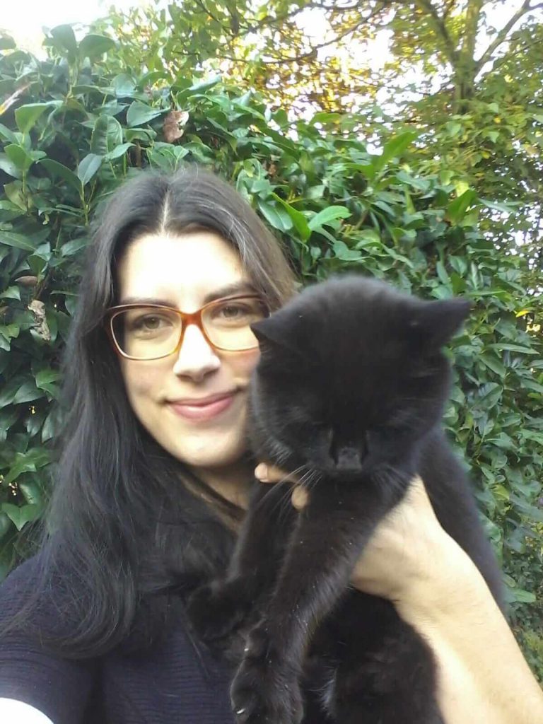 Eine Frau mit langen, schwarzen Haaren und Brille hält eine schwarze Katze im Arm. Im Hintergrund sind grüne Pflanzen sichtbar. Die Frau trägt ein dunkles Oberteil.